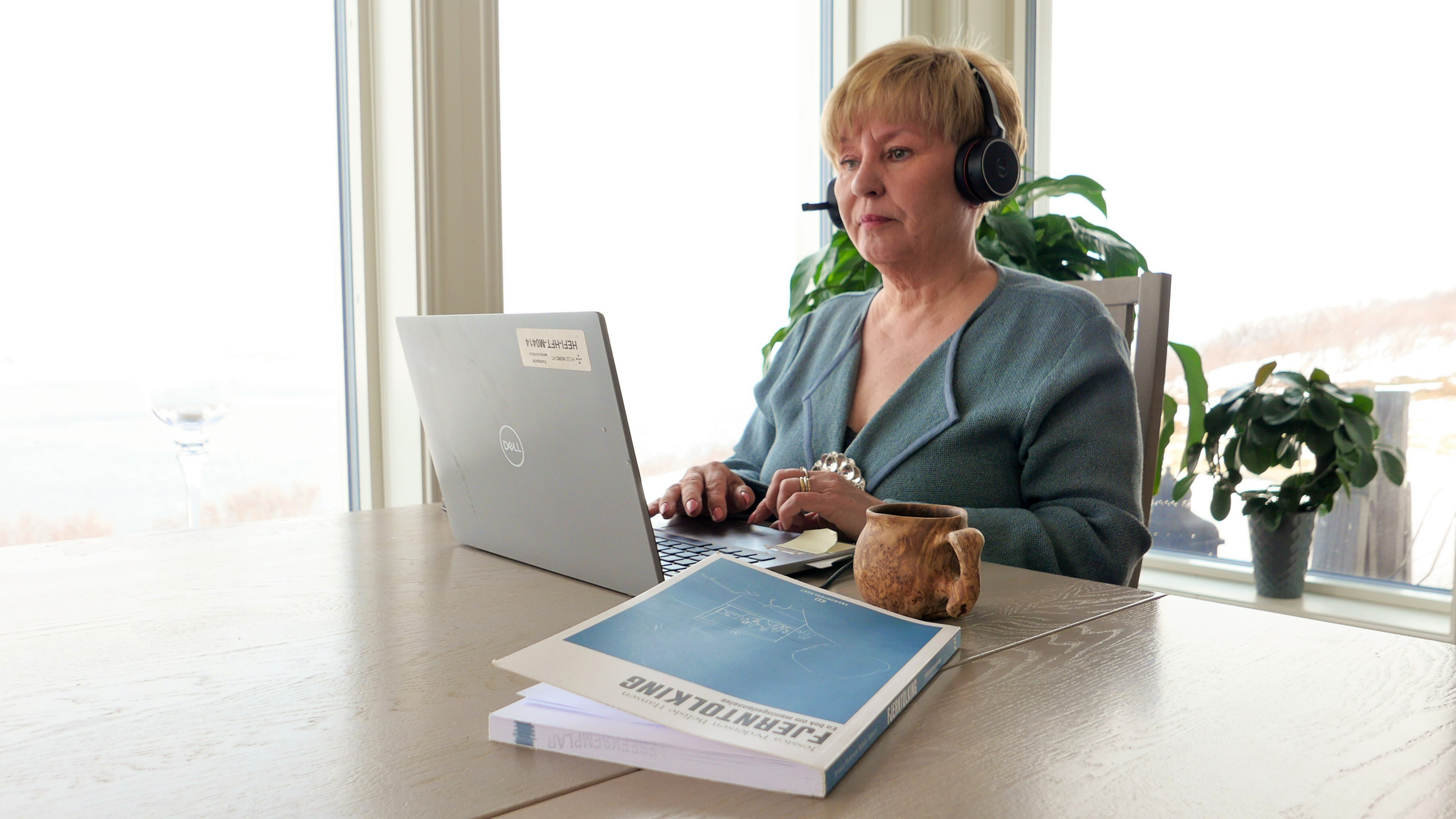 Kvinne sitter ved et spisebord og jobber med sin laptop, natur i bakgrunnen.
