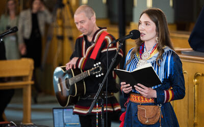 Kvinne og mann i samiske folkedrakter fremfører en vise, han spiller gitar og hun synger.