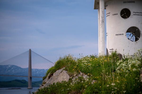 Hvitmalt betongskulptur med frodig gress og blomster i forkant. Ei bro i bakgrunnen.