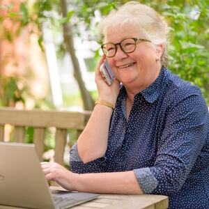 Eldre dame sitter på benk med PC og telefon.