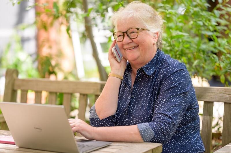 Eldre dame sitter på benk med PC og telefon.