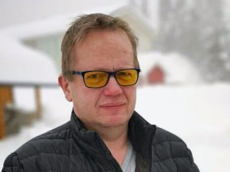 Pål Kjetil Lyngstad ser inn i kamera. Han har briller med gule glass, han har på seg grå genser under jakken. I bakgrunnen ser man snødekte hus og snødekte trær.