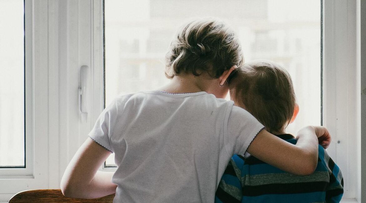 To barn som ser ut av et vindu. Det eldste barnet har armen sin beskyttende om det mindre barnet