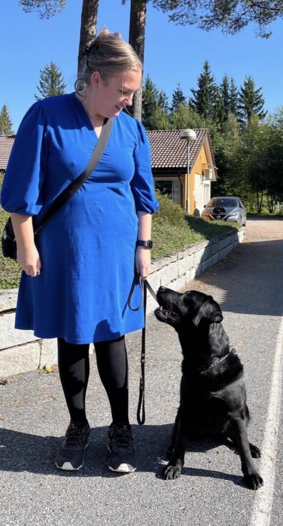 Dame i blå kjole står utendørs og ser på en svart hund som ser på henne.