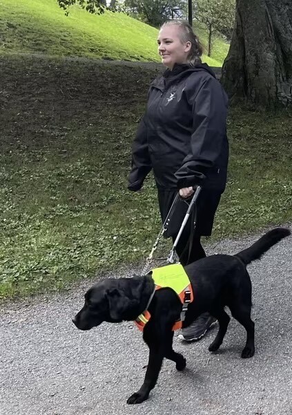 Kvinne med svart jakke går og holder en førerhundsele som er satt på en svart førerhund som går ved hennes side.