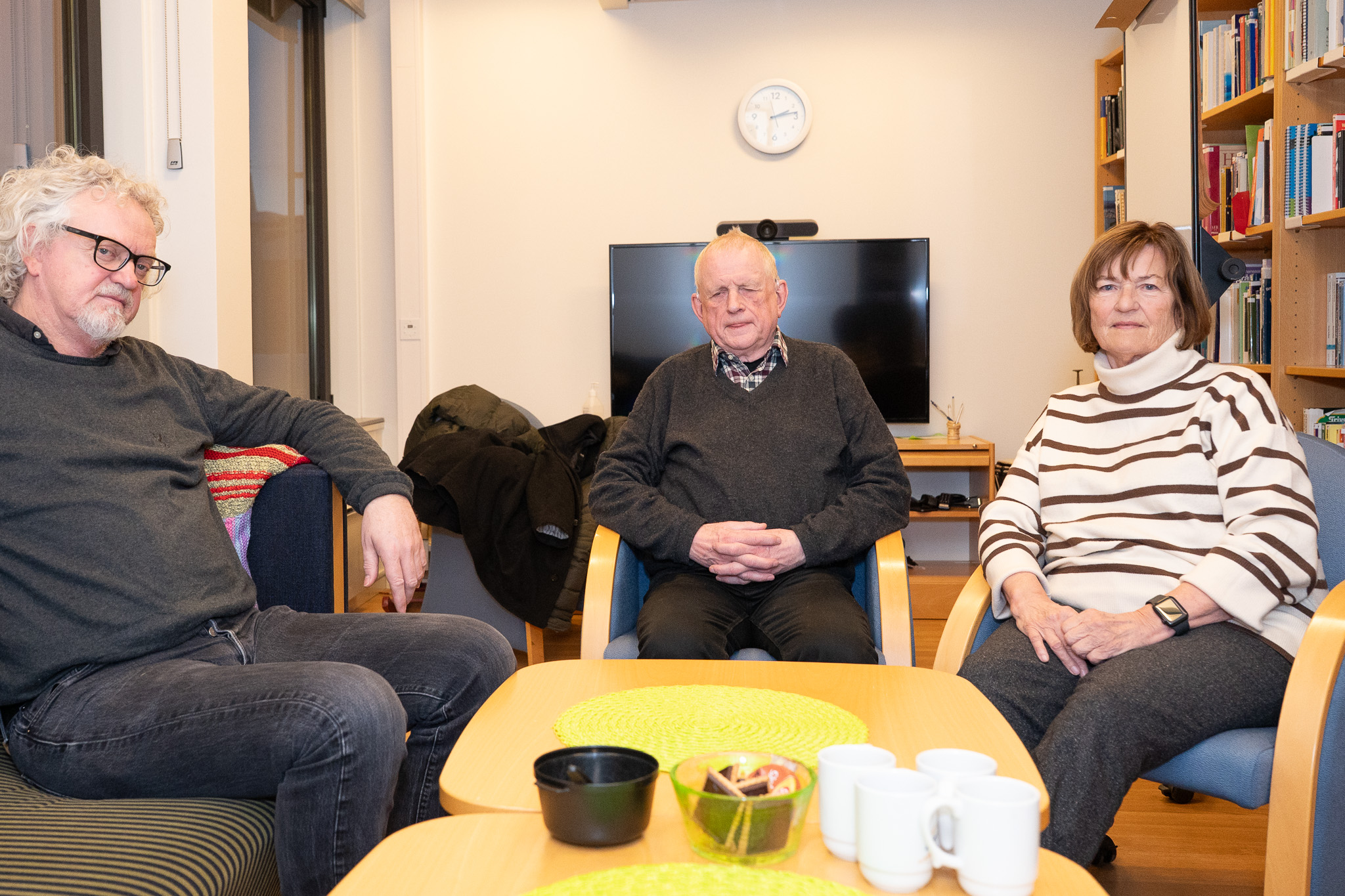 Tre personer sittende hvor alle ser mot kameraet. I bakgrunnen en TV-skjerm.