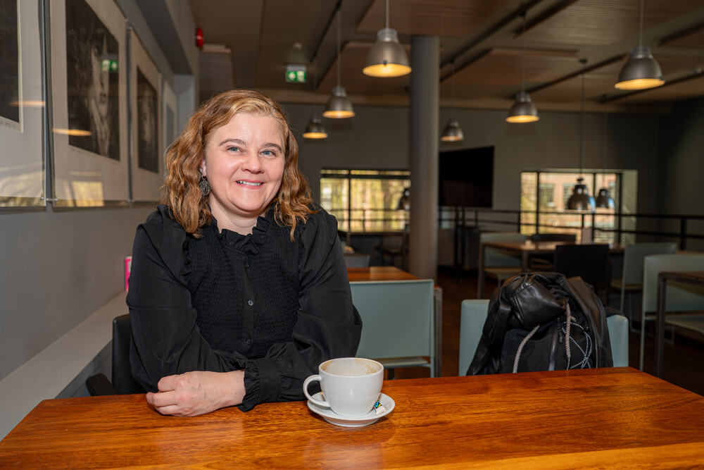 Kvinne med brunt hår med svart trøye sitter ved et bord med en hvit kopp foran henne.